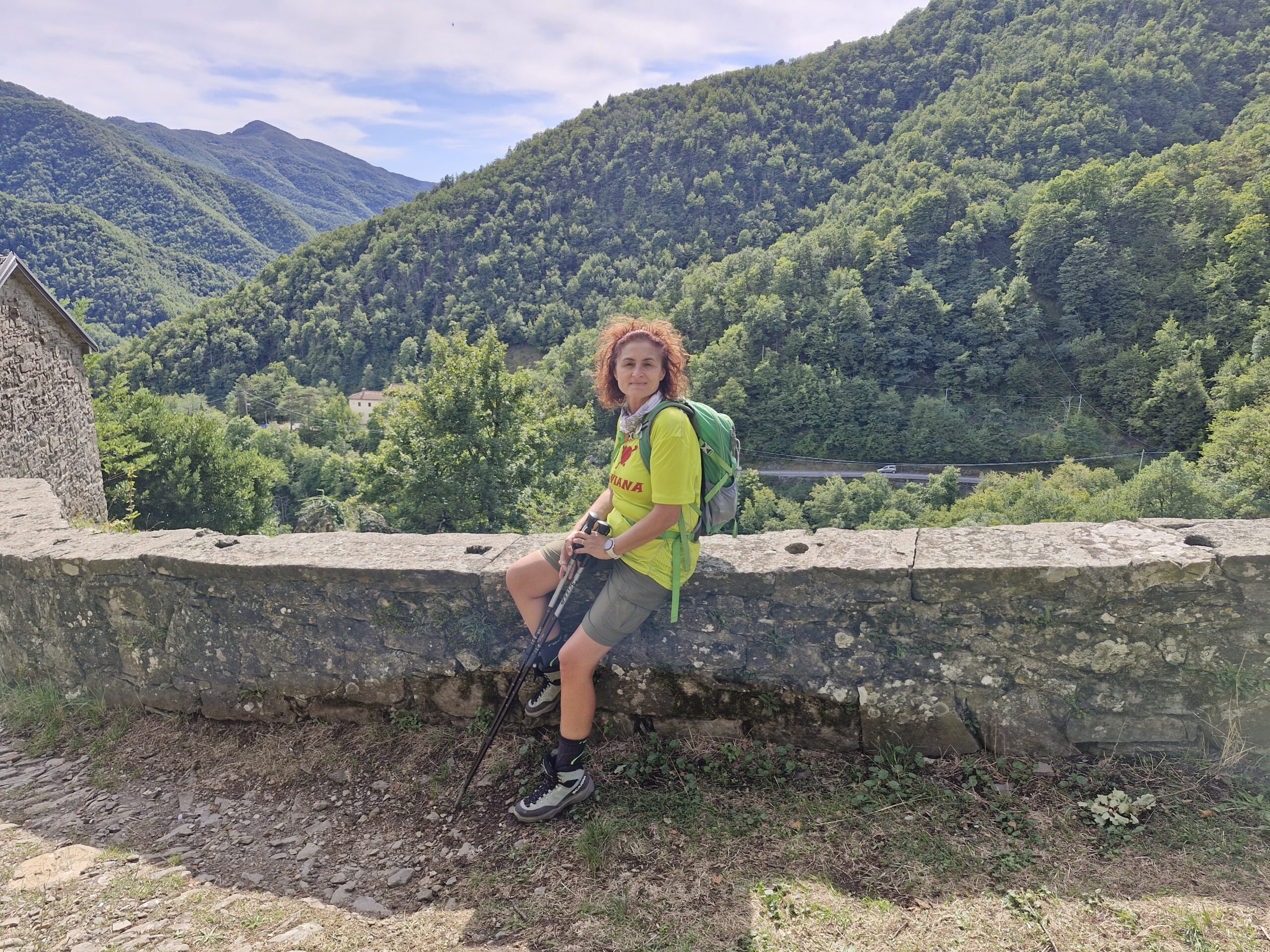 Una donna seduta su un muretto di pietra. Indossa una maglietta gialla, pantaloni corti e uno zaino verde. Ha due bastoncini da trekking e sorride. Lo sfondo è costituito da una collina verdeggiante. I migliori consigli di trekking