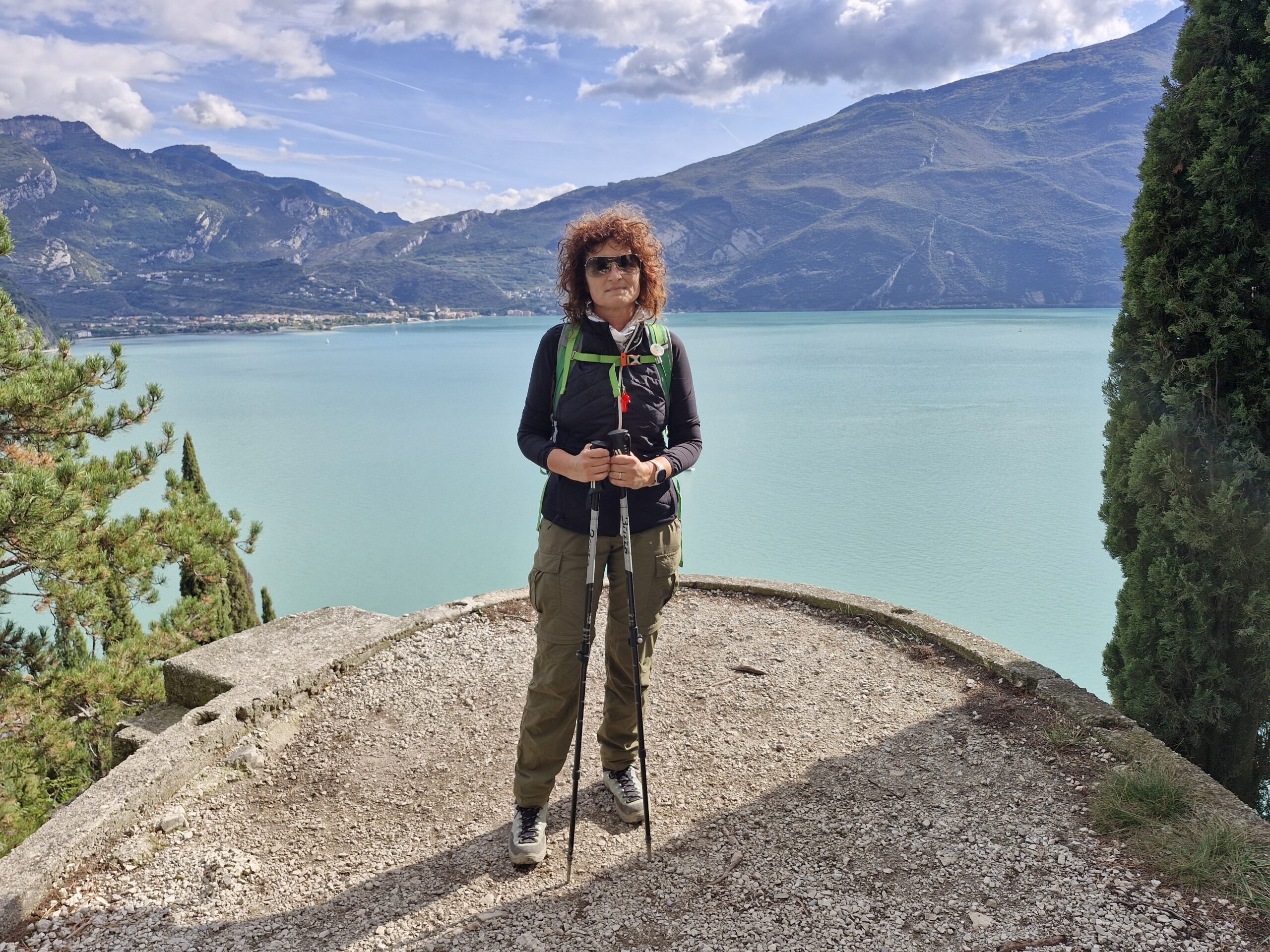 Una donna vestita da trekking su piattaforma di cemento sopra un lago di montagna di colore verdeacqua con colline verdi sullo sfondo. Il cielo è sereno e la scena anche.