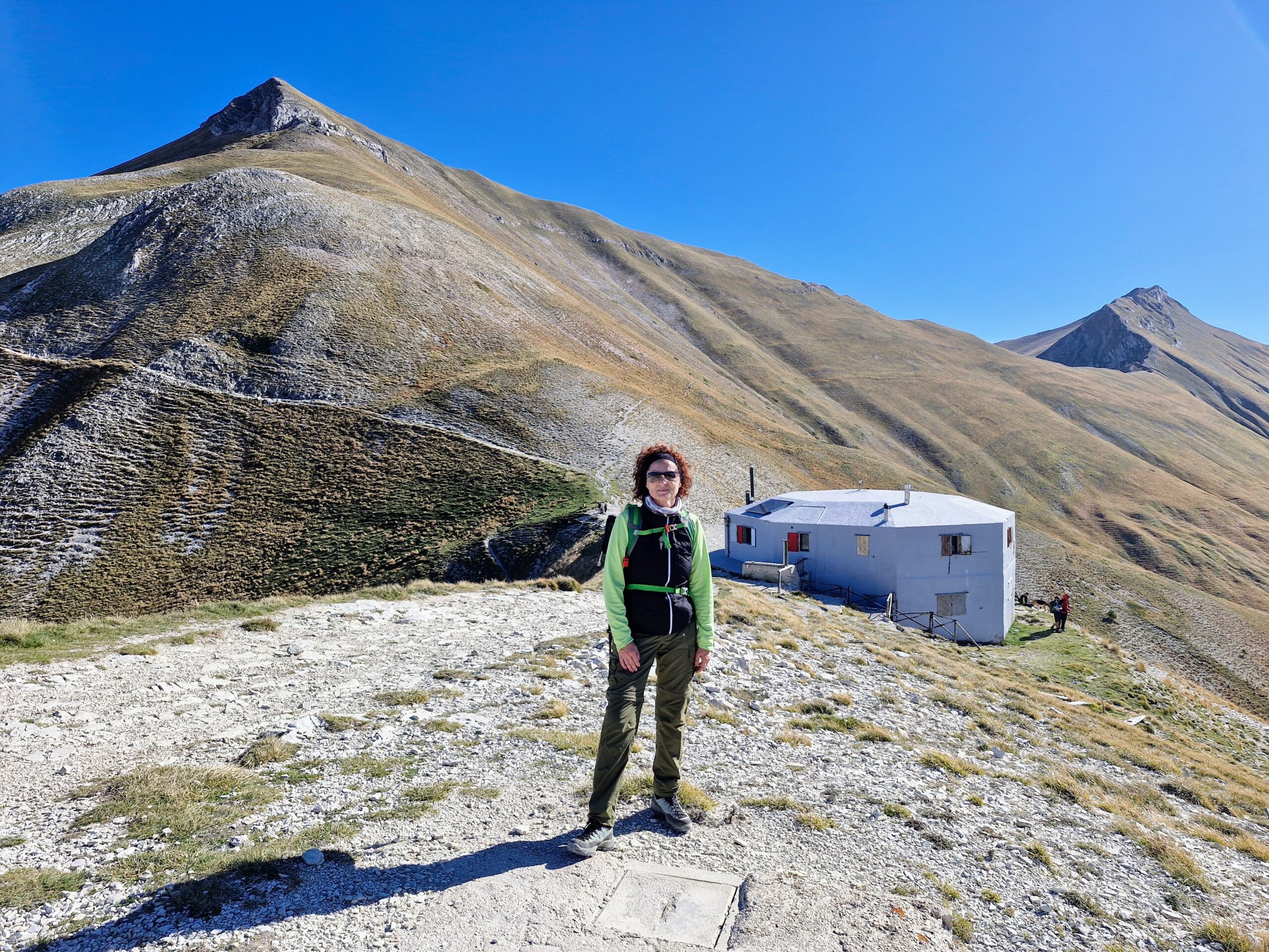 Donna vestita da trekking in montagna con cielo azzurro ed alle spalle un rifugio. I migliori consigli di trekking